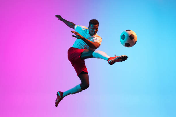 One African man, professional soccer football player training isolated on gradient blue pink background in neon light. Jumping, flying. One young African man, professional soccer football player training isolated on gradient blue pink background in neon light. Concept of action, energy, sport. Copy space for ad match lighting equipment photos stock pictures, royalty-free photos & images