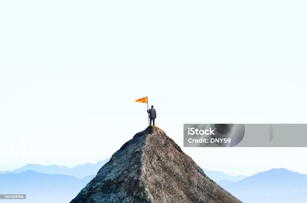 Businessman At Top Of Mountain Peak Holds Large Flag A businessman stands at the top of a mountain peak as he holds a large orange flag attached to a pole. He stands with his back to the camera as he looks out into the distance towards a distant mountain range that sits on the horizon. Aspirations Stock Photo