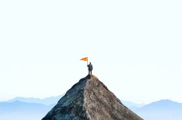 Businessman At Top Of Mountain Peak Holds Large Flag A businessman stands at the top of a mountain peak as he holds a large orange flag attached to a pole. He stands with his back to the camera as he looks out into the distance towards a distant mountain range that sits on the horizon. mountain peak stock pictures, royalty-free photos & images