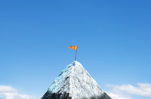 An orange flag is planted on top of a snow cap peak. Soft clouds frame the lower portion of the image and a rich blue sky provides ample room for copy or text.