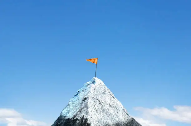 An orange flag is planted on top of a snow cap peak. Soft clouds frame the lower portion of the image and a rich blue sky provides ample room for copy or text.