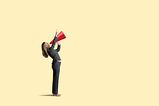 A woman standing with a red megaphone points in the direction she is shouting as she attempts to get someone'satention. Isolated on a yellow background.
