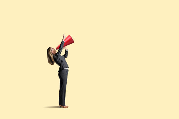 Woman Shouting Through Megaphone A woman standing with a red megaphone points in the direction she is shouting as she attempts to get someone'satention. Isolated on a yellow background. beckoning photos stock pictures, royalty-free photos & images