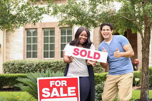 Mixed race family of African and Latin descent buys their first home.  Real estate sign and home in background.  Spring or summer season.