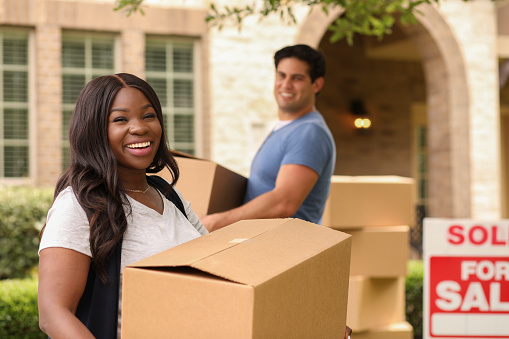 Mixed race family of African and Latin descent moving boxes into a new home.  Real estate sign and home in background.  Spring or summer season.