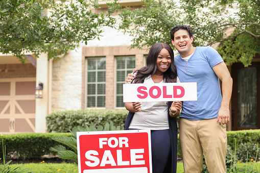 Mixed race family of African and Latin descent buys their first home.  Real estate sign and home in background.  Spring or summer season.