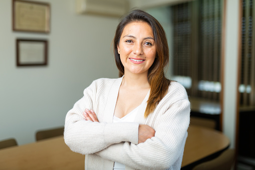 Young confident hispanic businesswoman posing with crossed arms in modern office interior, smiling at camera