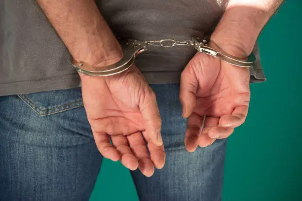 Photo of Arrested man handcuffed hands at the back. Cropped view of handcuffed man in denim jeans standing isolated on green, human rights concept