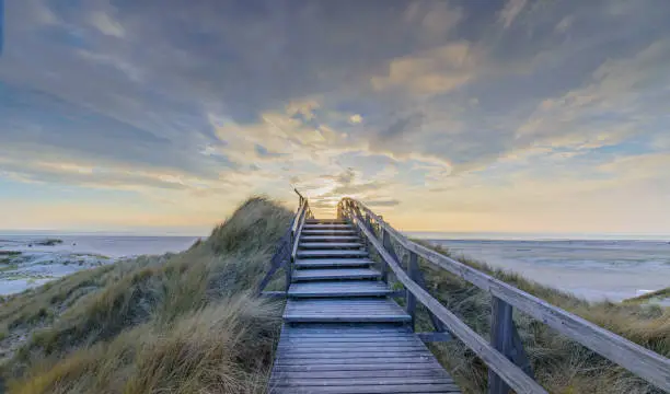Photo of Boardwalk and stairs leading through the dunes to the beach of North Sea by sunset in Norddorf, Amrum, Schleswig-Holstein. Bathing and beach vacation on the North Sea.
