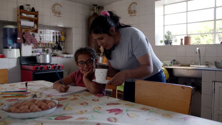 Loving mom offering hot chocolate to daughter while supervising her homework both looking happy