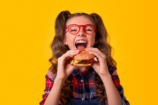 Funny hungry girl in casual clothes and glasses with mouth opened biting hamburger and looking at camera against yellow background