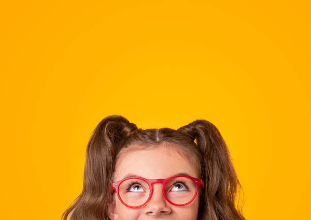 Happy girl in trendy glasses looking up Positive teen girl with ponytails wearing stylish red framed eyeglasses looking up on yellow background with empty space looking up stock pictures, royalty-free photos & images