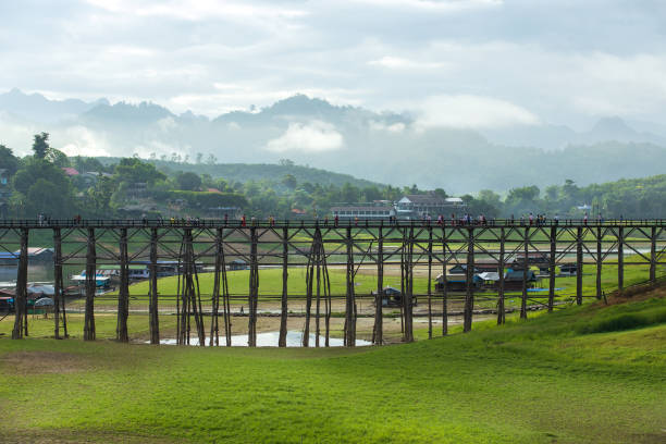 vue de l’ancien pont en bois (pont mon) et de la rivière avec du brouillard sur les montagnes et des touristes marchant sur le pont le matin à sangkhlaburi, province de kanchanaburi, thaïlande. - asia kanchanaburi province lake nature photos et images de collection