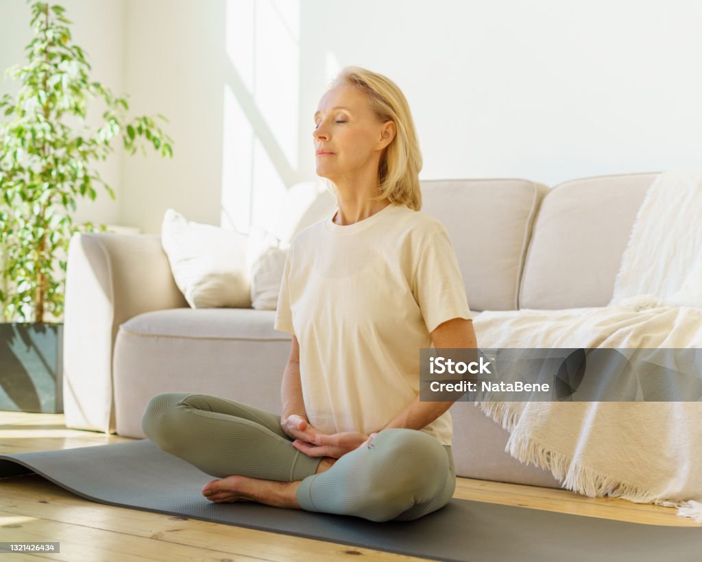 Retired woman meditating and practicing yoga while sitting in lotus pose on floor at home Peaceful senior woman in lotus position meditation with closed eyes at home while sitting on yoga mat on floor, full length. Calm elderly lady practicing meditation techniques and yoga indoors Meditating Stock Photo