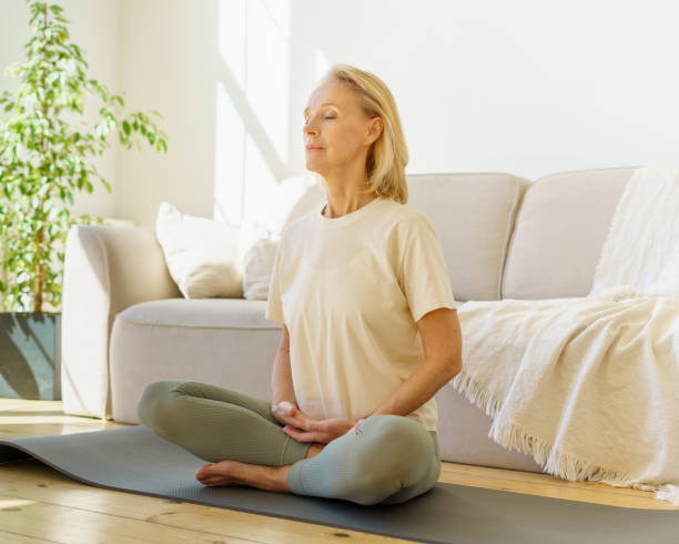 Retired woman meditating and practicing yoga while sitting in lotus pose on floor at home Peaceful senior woman in lotus position meditation with closed eyes at home while sitting on yoga mat on floor, full length. Calm elderly lady practicing meditation techniques and yoga indoors meditating stock pictures, royalty-free photos & images