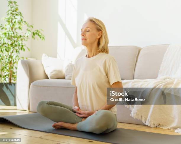 Retired Woman Meditating And Practicing Yoga While Sitting In Lotus Pose On Floor At Home Stock Photo - Download Image Now