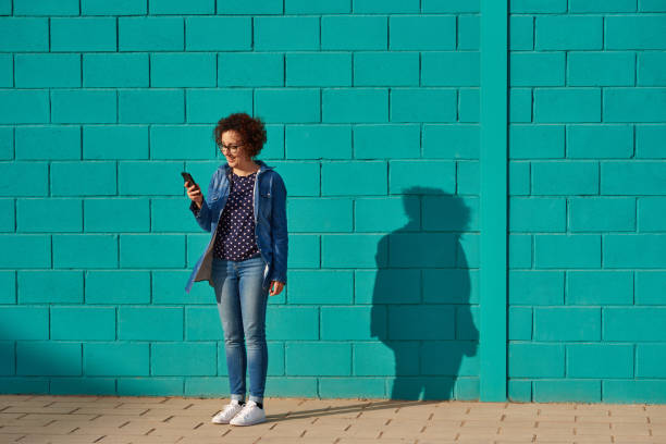 istock Front view of a happy woman standing against a bright blue wall using her smartphone 1321426718