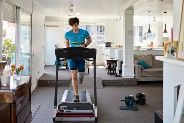 Full length front view of Caucasian man with prosthetic leg running on exercise machine in living room of family home.