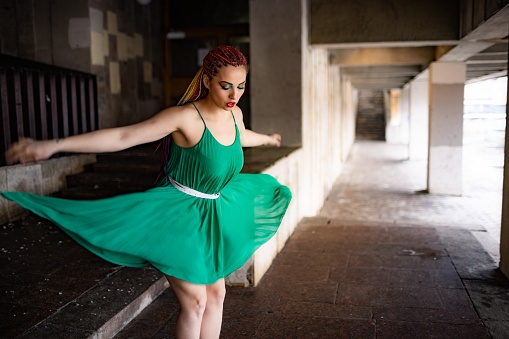 A girl with a surprised face with bright African braids and rainbow glitter makeup in a light green spring dress. Jumping off the steps of a large old building in a warm spring city.
