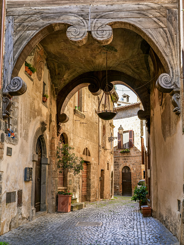 A suggestive view of an ancient and picturesque alley with tuff houses in the heart of the medieval town of Orvieto in the Umbria region, in central Italy. The decoration of the upper arch is completely in inlaid solid wood. With a population of just 20,000 people, Orvieto is considered one of the most beautiful cities of art in Italy, founded since the Etruscan and Roman times on the flat top of a large butte of volcanic tuff. This same material was used for the construction of almost all the medieval houses and churches of Orvieto, as well as its famous Gothic-style Cathedral. Image in high definition format.