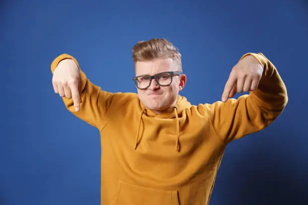 Young handsome man dressed in casual yellow sweater and glasses on a blue background, Surprised pointing down with his finger, with an open mouth and an amazed expression on his face. By encouraging subscribers to subscribe or follow the link in the description.