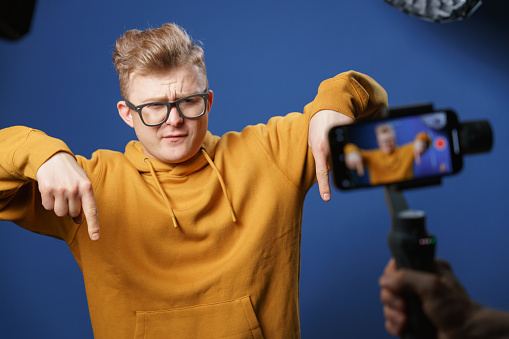 The vlogger shoots a new video for his subscribers on his smartphone. The blogger points his fingers down, with this gesture he asks new viewers to subscribe to the channel, as well as to like and leave a comment. The vlogger records a new video where he comments on new products from the world of modern technologies.