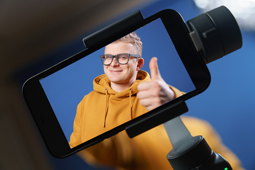 Everything is cool! Everything is fine! The vlogger shows a thumbs-up gesture, in front of a smartphone that is shooting a video. A close-up photo of the smartphone that takes it. Good mood and the mood for success.