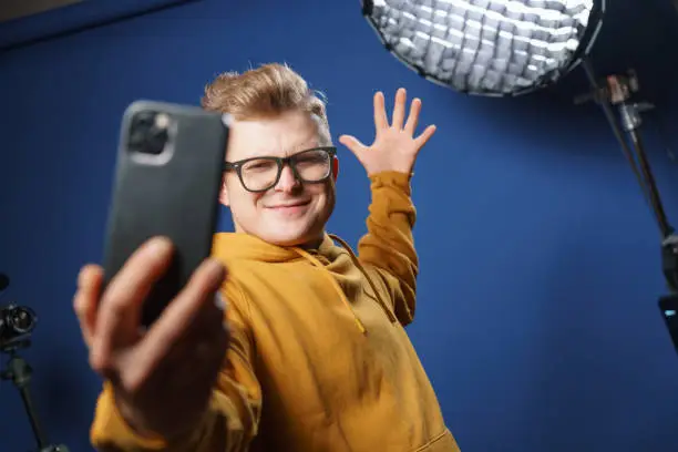 Photo of A vlogger of 30 years old in a yellow hoodie and glasses, very positively takes a selfie on his expensive and modern smartphone.