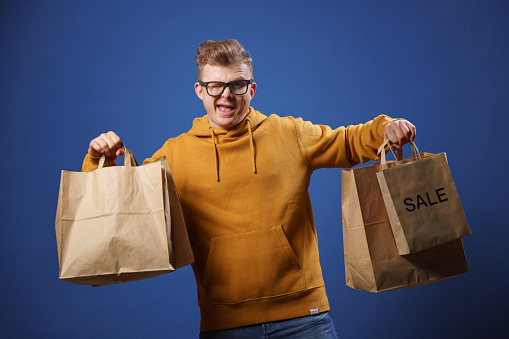 A photo of a cheerful, funny, man in a hoodie and glasses who is holding paper bags with purchases made in an online store. Make purchases in online stores, easily and quickly. Save your money on sales. Shopping without leaving home. The delivery service will deliver your order quickly and on time.