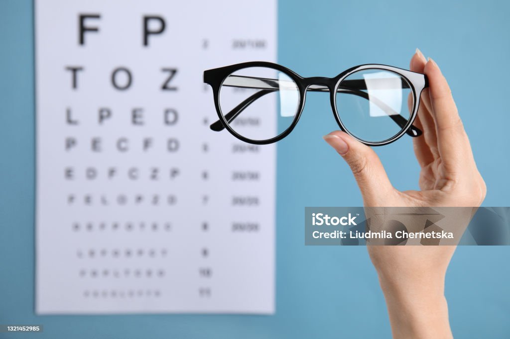 Woman holding glasses against eye chart on blue background, closeup. Ophthalmologist prescription Eyeglasses Stock Photo