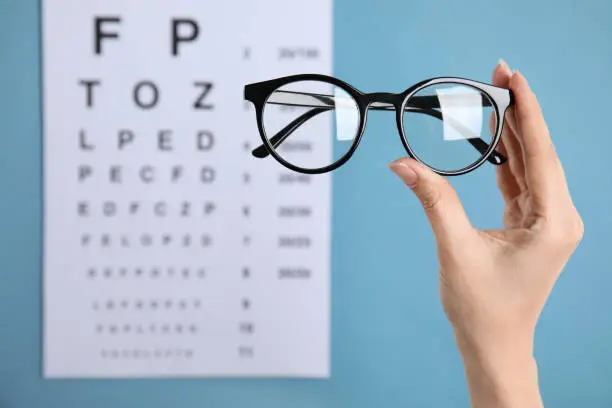 Photo of Woman holding glasses against eye chart on blue background, closeup. Ophthalmologist prescription