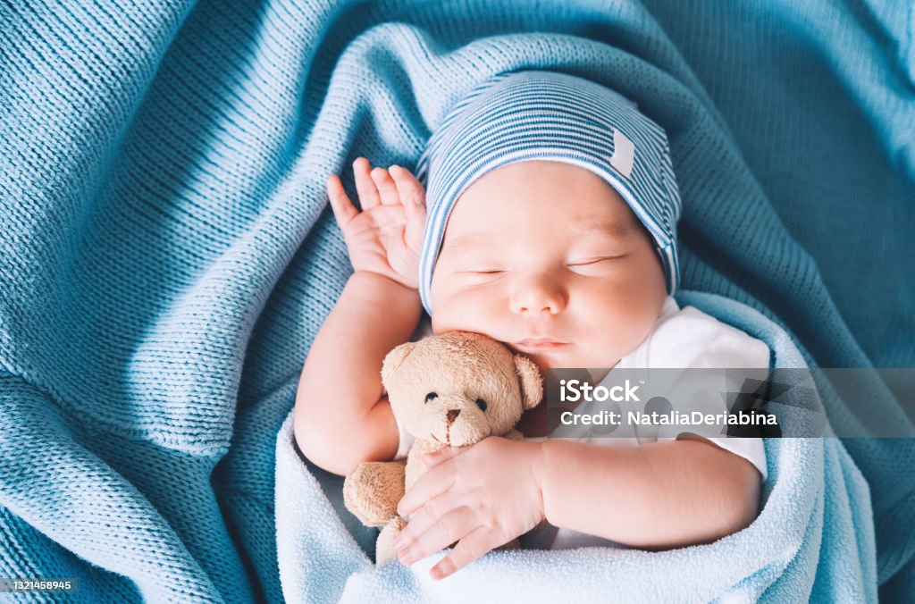 Newborn baby sleep at first days of life. Portrait of new born child boy one week old sleeping peacefully with a cute soft toy in crib in cloth background. Baby - Human Age Stock Photo