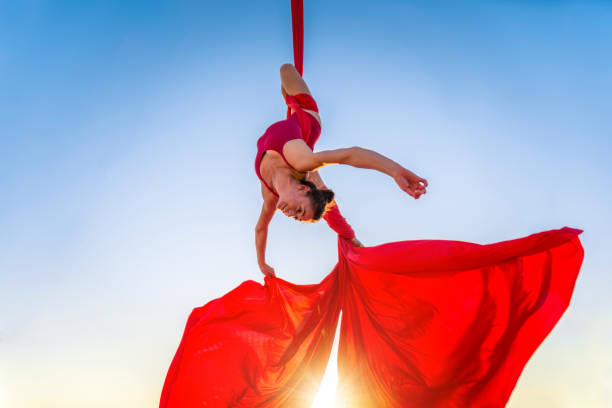 athletische, akrobat turner durchführung luftübung mit roten stoffen im freien auf himmel hintergrund. flexible frau im roten anzug führt zirkuskünstler tanzen in der luft auf seide auf dem kopf. sonnenlicht - akrobat stock-fotos und bilder