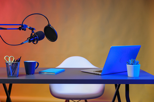 A recording video studio with a modern gray table on metal legs, a black microphone with arm stand on it and a thin silver laptop, a pot with a flower and a notebook