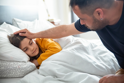 Dad worrying about his daughters health sitting on bed. Sick and tired girl lying on bed covered with blanket. Caucasian man checking kids forehead. Fatherhood, healthcare concept