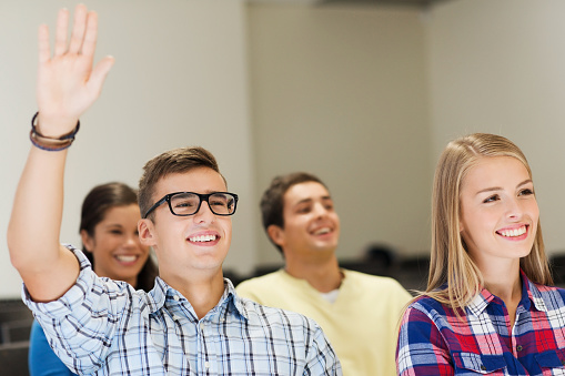 education, high school, teamwork and people concept - group of smiling students raising hand in lecture hall
