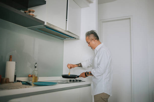 homme âgé chinois asiatique brisant œuf de friture avec des casseroles de cuisson dans la cuisine pendant la matinée se préparant pour le petit déjeuner - break eggs domestic kitchen breaking photos et images de collection