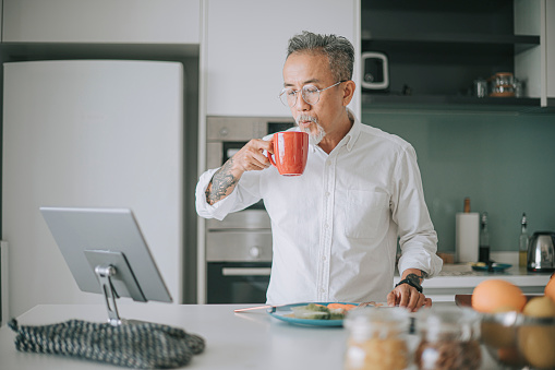 Asian chinese gray hair senior man with hair stubble enjoying breakfast at kitchen counter weekend while using digital tablet