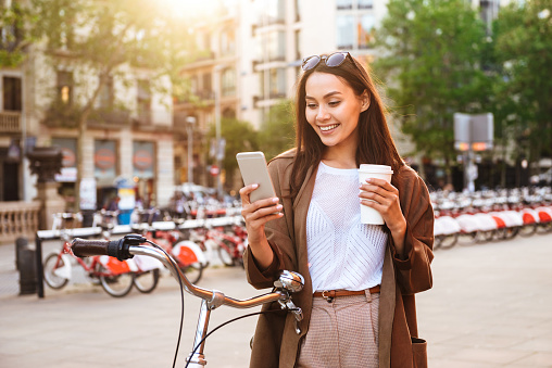 Image of young woman outdoors with bicycle on the street looking aside chatting by mobile phone drinking coffee.