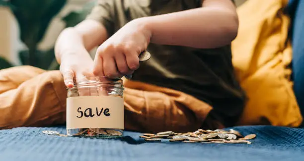 Photo of Close up of Little child kid boy hands grabbing and putting stack coins in to glass jar with save label. Donation, saving money, charity, family finance plan concept