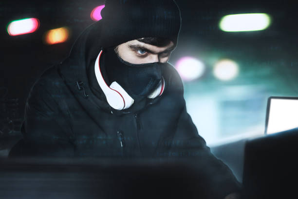 istock Angry computer hacker weared balaclava stealing data from laptop in front of black background and blue light. close portrait. 1321475172