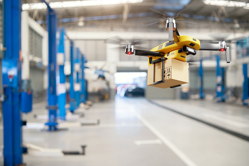 Flying delivery drone transferring parcel box from distribution warehouse to automotive garage customer service repair center background. Modern innovative technology and gadget concept.