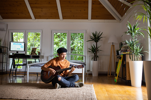 Japanese man sitting on floor and enjoying while playing acoustic guitar in bedroom at home