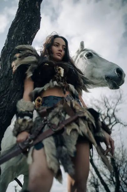 Photo of Woman stands in forest in image of warrior amazon with sword near her horse.