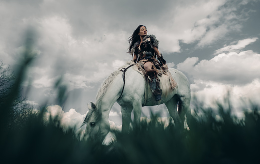 Woman rides on horseback in field in image of ancient warrior amazon on sky background.