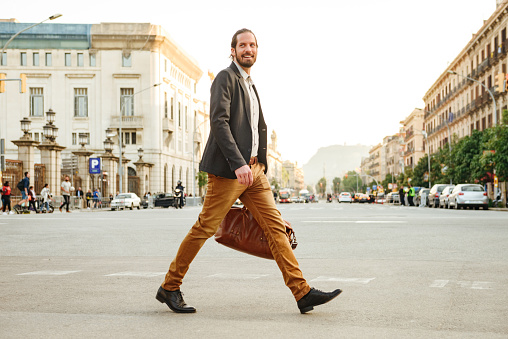 Portrait of elegant businessman in jacket laughing while crossing road in megapolis with leather male bag in hand