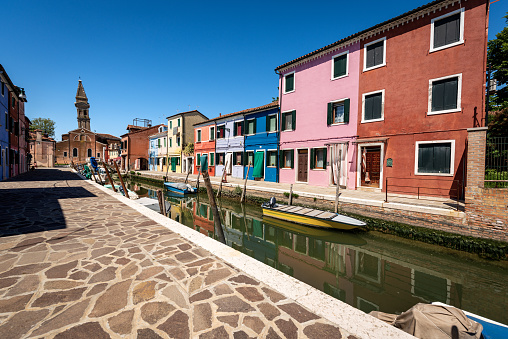 Multi colored houses and moored small boats along a canal in Burano island, Venice lagoon, UNESCO world heritage site, Veneto, Italy, Europe. Church of San Martino with leaning bell tower.