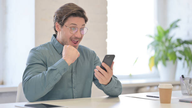 istock Middle Aged Man Celebrating Success on Smartphone in Office 1321491049