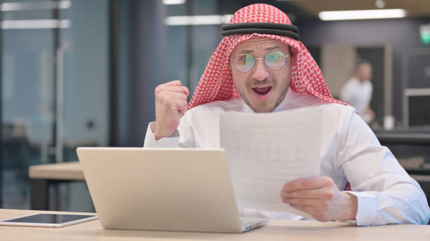 Middle Aged Man with Laptop Celebrating Success on Documents The Middle Aged Man with Laptop Celebrating Success on Documents middle eastern ethnicity mature adult book reading stock pictures, royalty-free photos & images