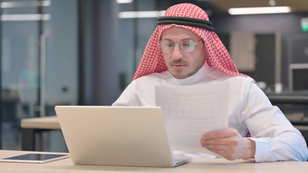 Middle Aged Man with Laptop Reading Documents The Middle Aged Man with Laptop Reading Documents middle eastern ethnicity mature adult book reading stock pictures, royalty-free photos & images